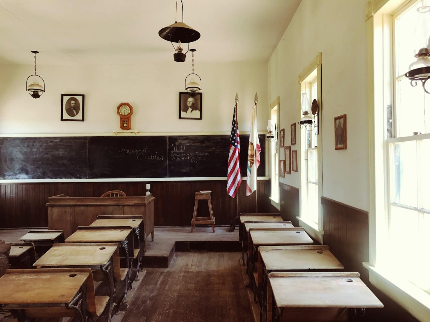 Sunlight streams into an empty old-fashioned classroom with wooden desks.