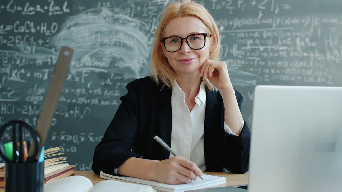 Woman in blazer and glasses smiles at desk with blackboard.
